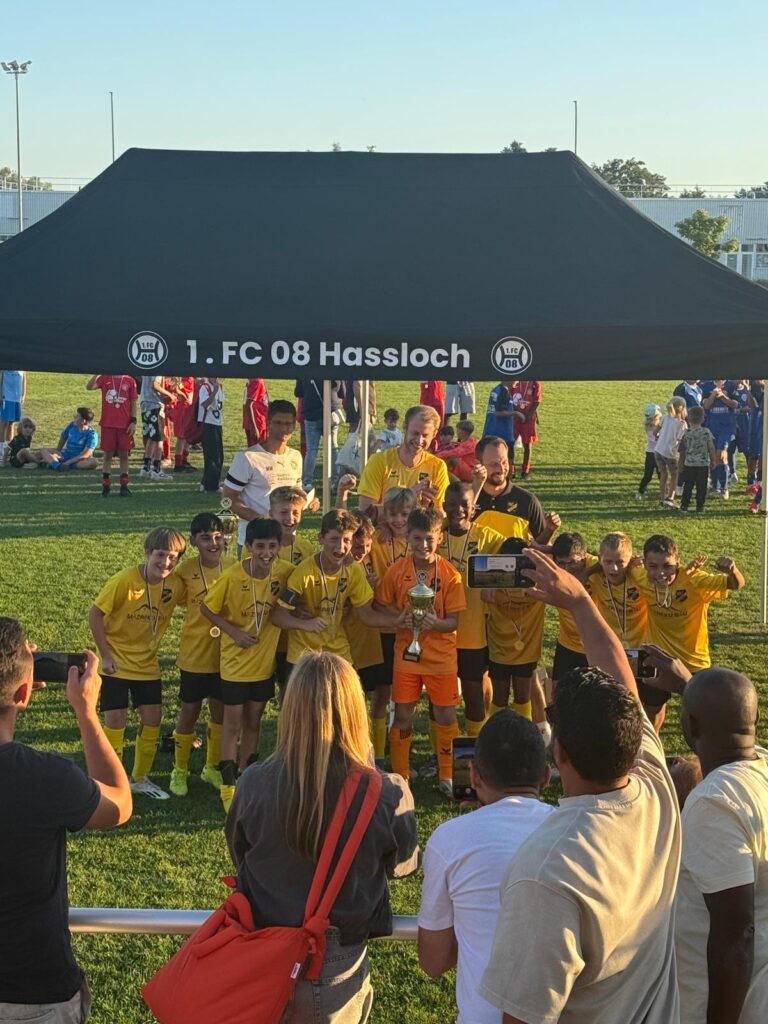 Gruppe von jungen Fußballspielern in gelben Trikots posiert mit Pokal unter einem Zelt.