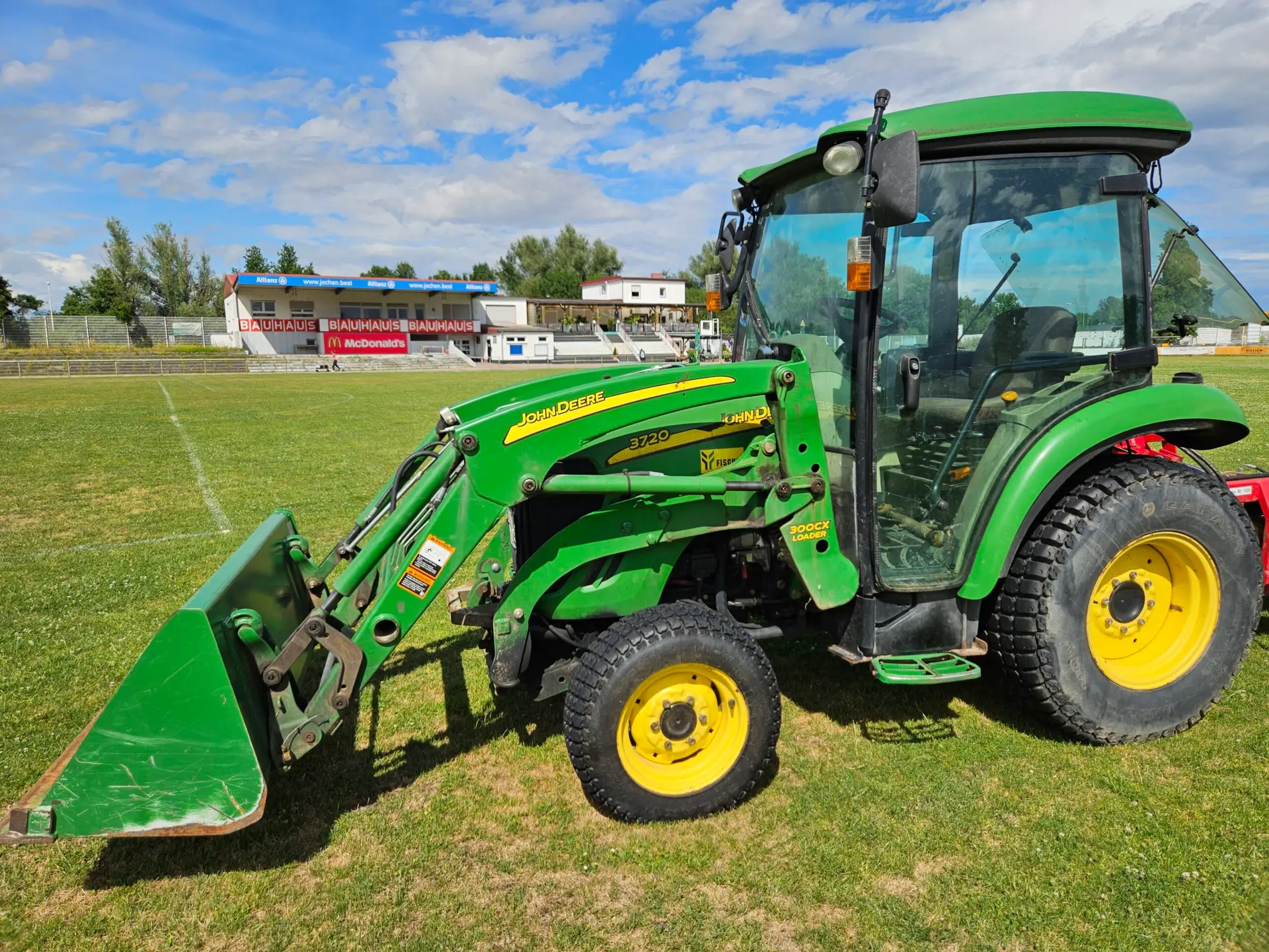 Grüner John Deere Traktor mit Frontlader auf einem Sportplatz.
