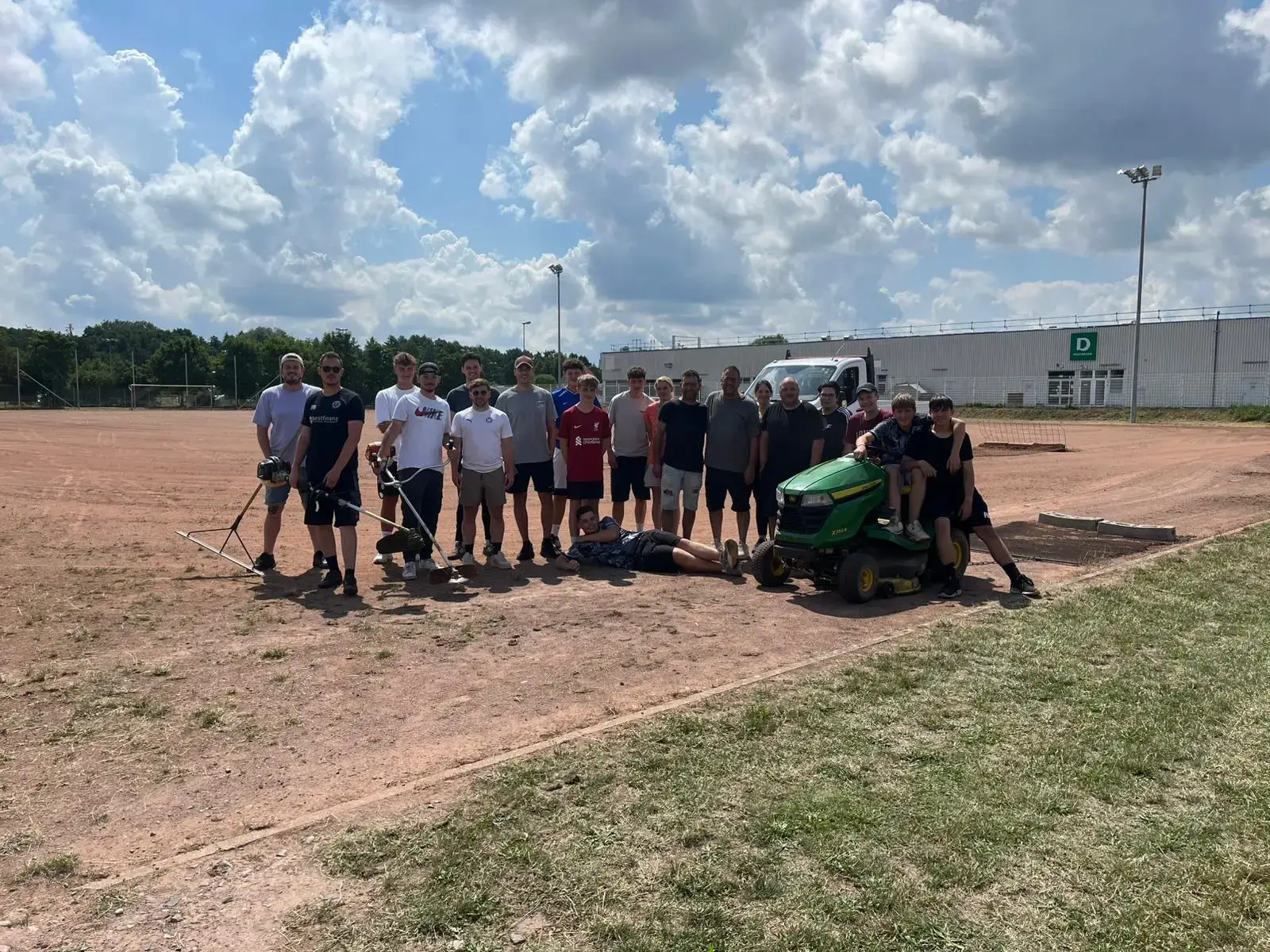 Gruppe von Personen auf einem Sportplatz, einige halten Werkzeuge, andere stehen oder sitzen auf dem Boden.