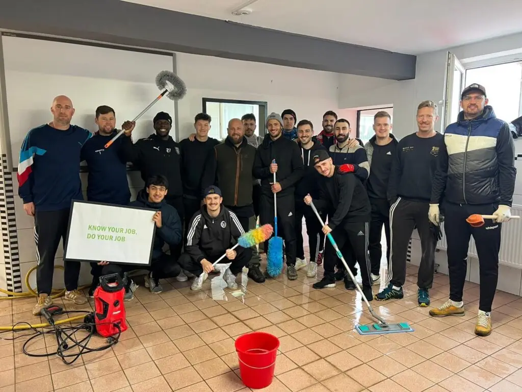 Fußballmannschaft posiert mit Reinigungsutensilien in einem Raum, einige halten Schilder.