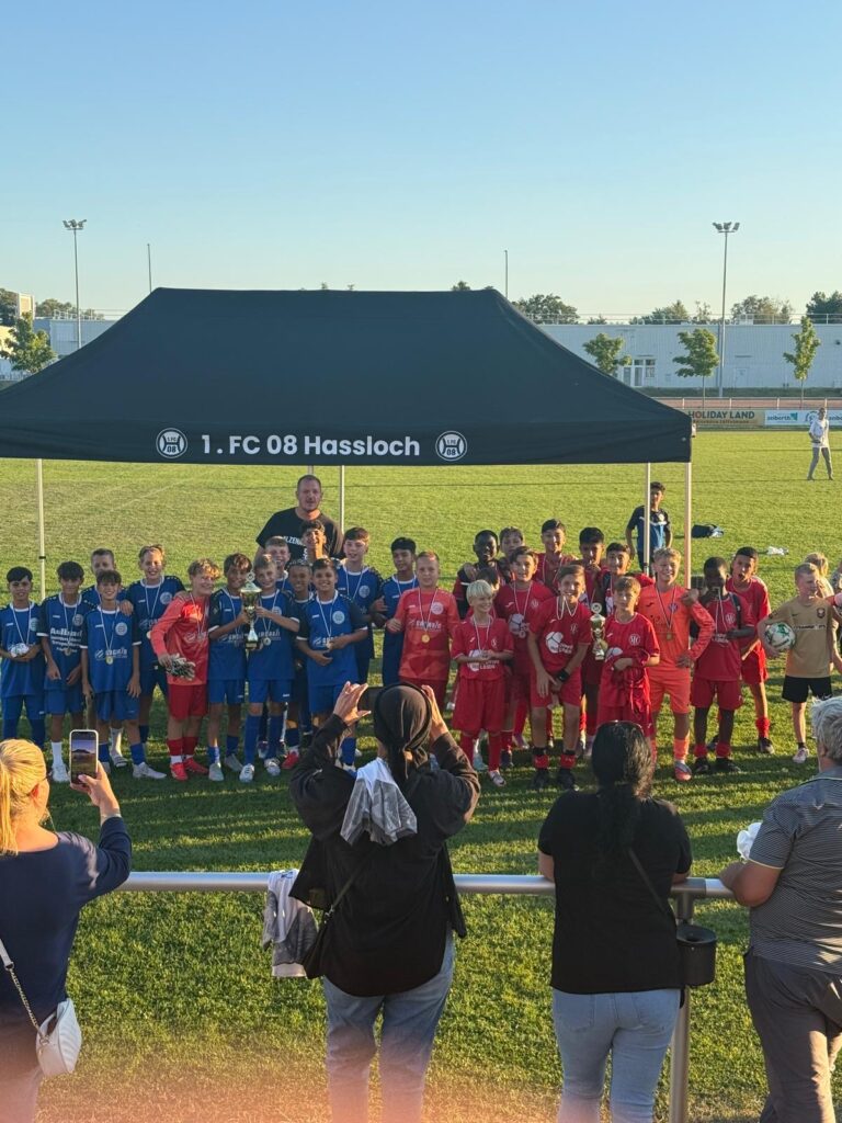 Zwei Fußballmannschaften in blauen und roten Trikots stehen unter einem Zelt mit Medaillen nach einem Turnier.