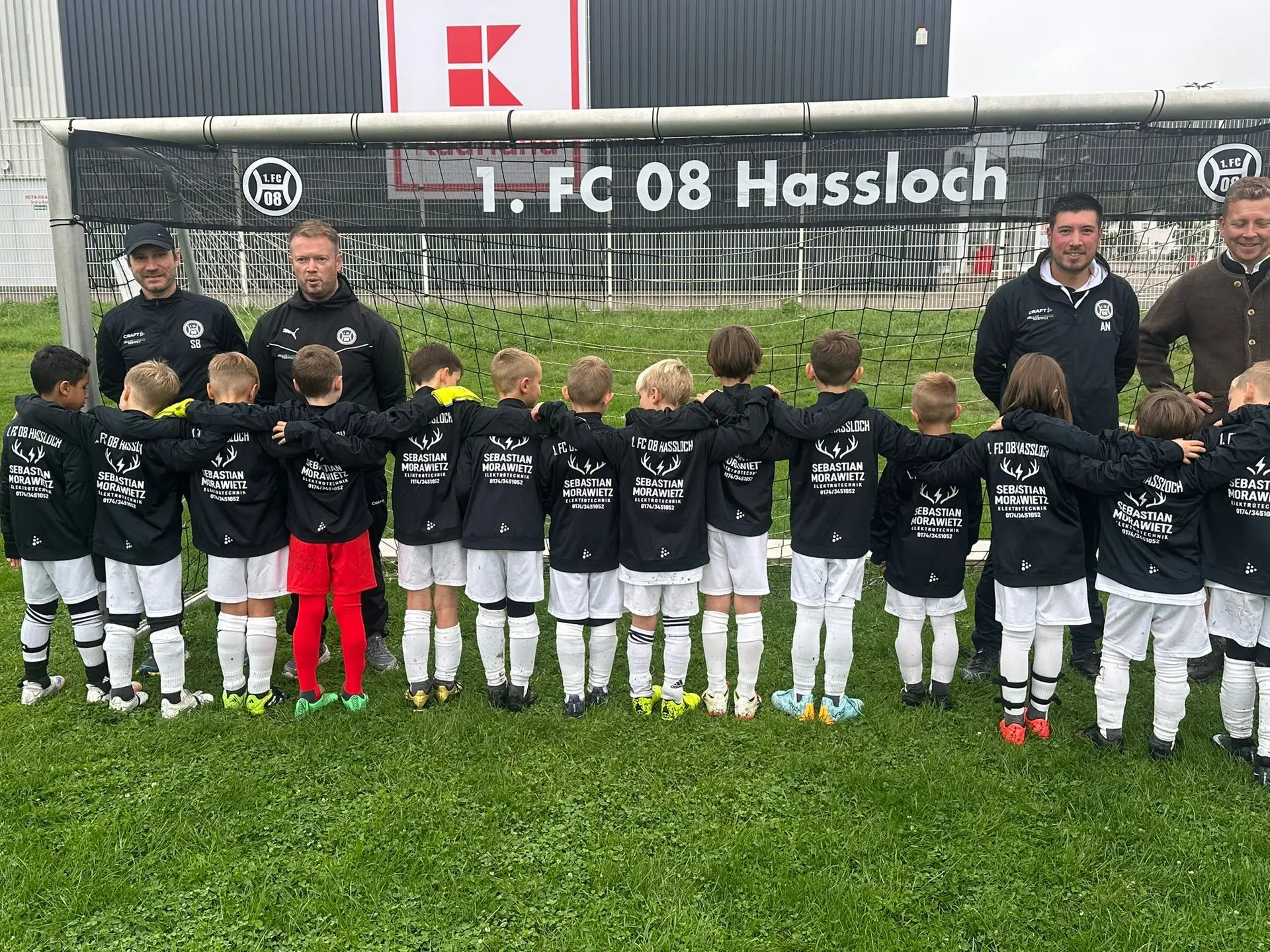 Gruppe von Kindern in schwarzen Trainingsjacken vor einem Fußballtor mit dem Logo von 1.FC 08 Hassloch.