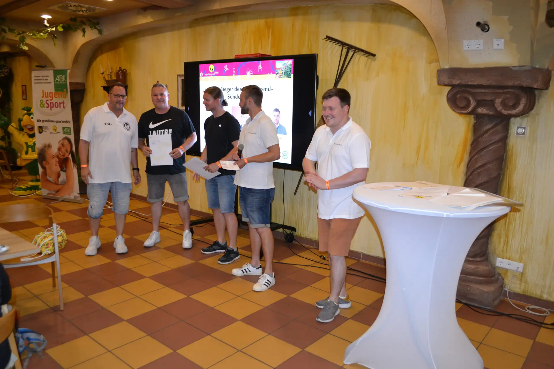 Fünf Personen stehen zusammen bei einer Veranstaltung, während eine Präsentation im Hintergrund zu sehen ist.