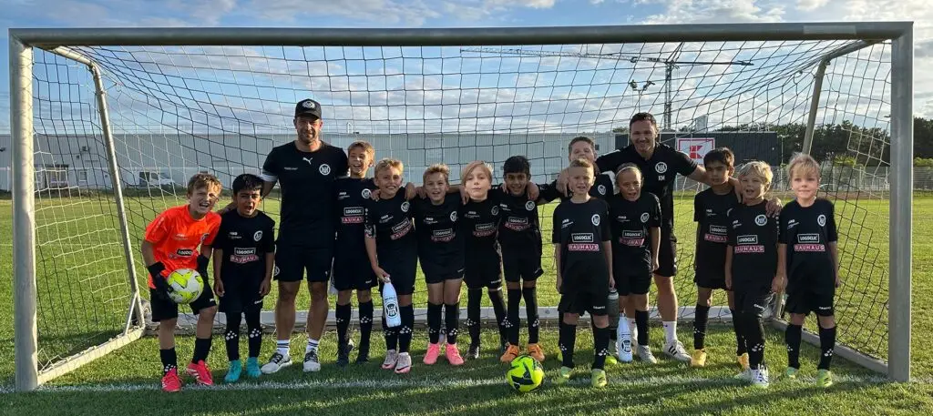 Eine Jugendfußballmannschaft in schwarzen Trikots steht mit Trainern vor einem Tor auf dem Spielfeld.