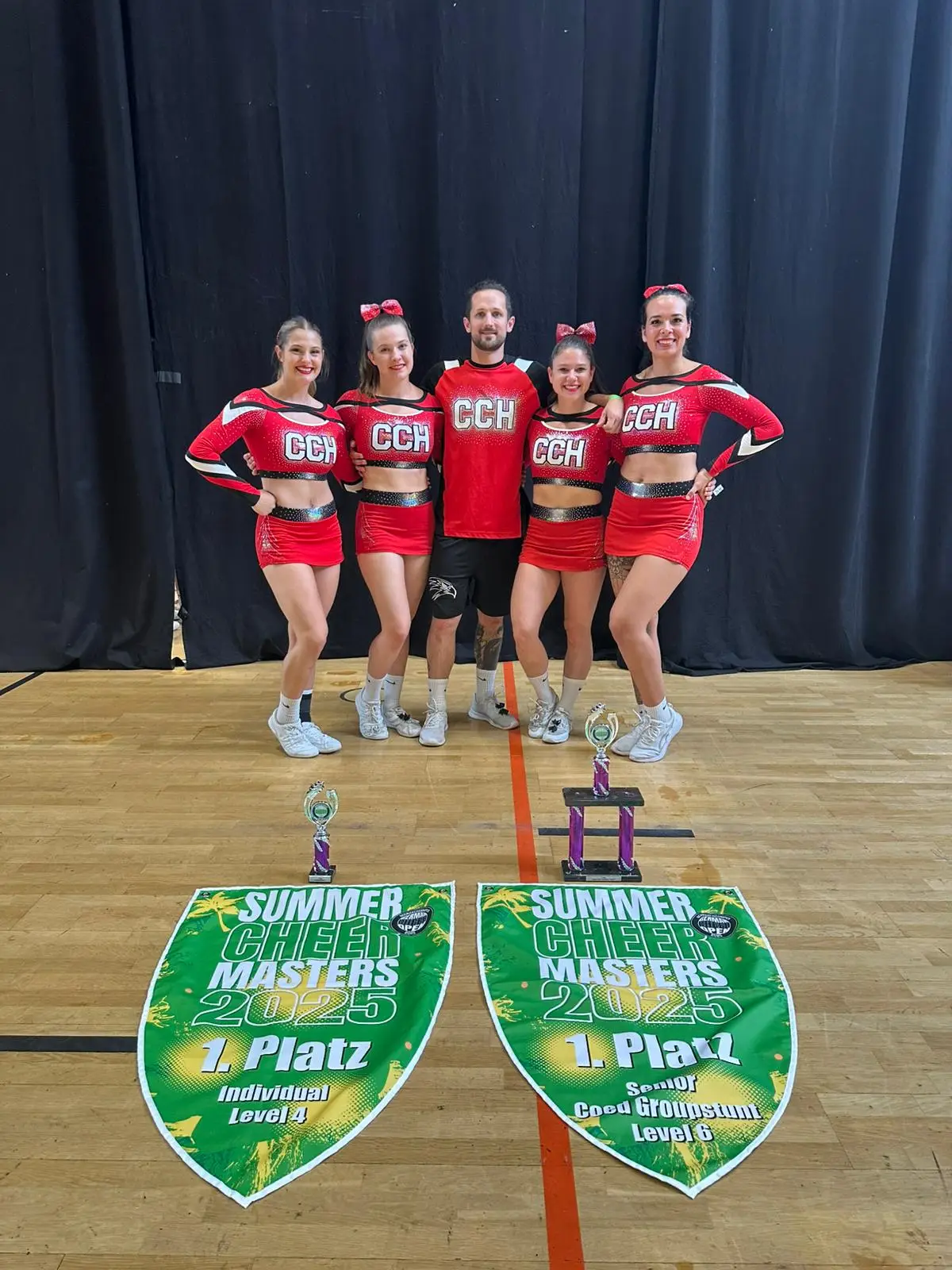 Cheerleading-Team mit roten Outfits und Trophäen auf dem Boden, die den ersten Platz zeigen.