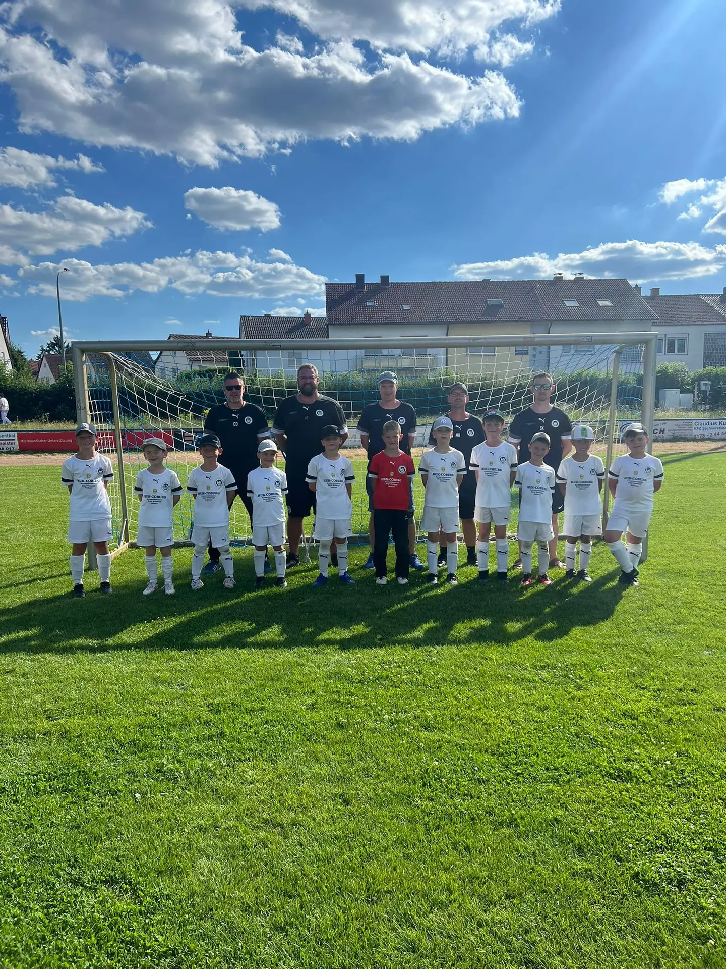 Eine Fußballmannschaft in weißen Trikots steht vor einem Tor auf dem Spielfeld mit Trainern im Hintergrund.