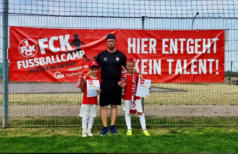 Zwei Kinder in Fußballkleidung mit einem Trainer vor einem Banner des FCK Fußballcamps.