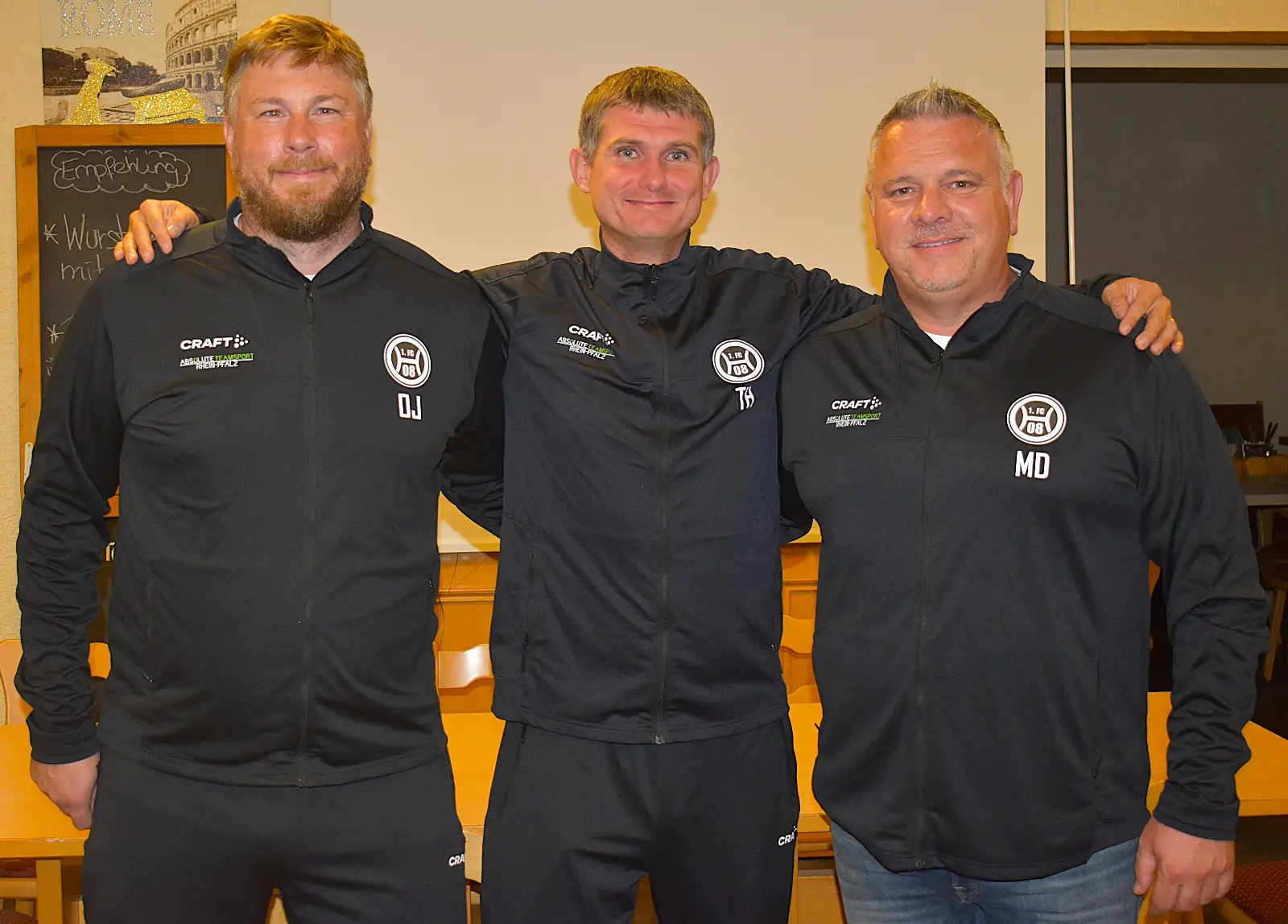 Drei Männer in schwarzen Trainingsjacken stehen nebeneinander und lächeln in die Kamera.