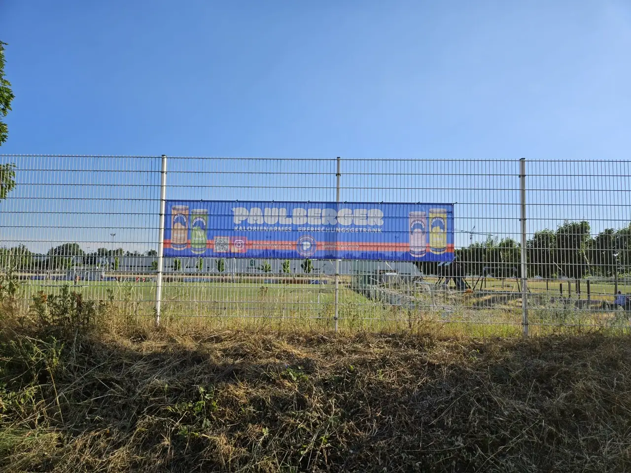 Ein großes Banner mit dem Paulaner Bierlogo, das an einem Zaun befestigt ist.