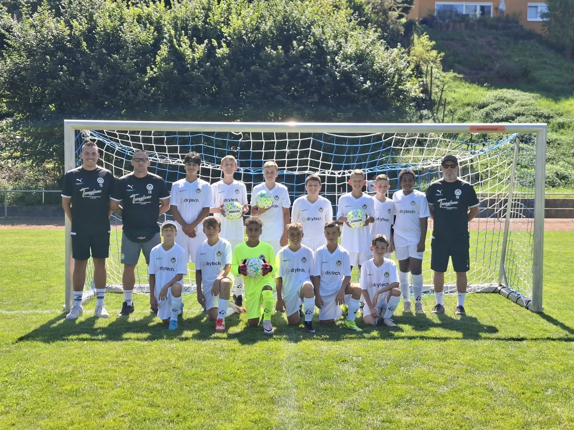 Eine Fußballmannschaft mit Trainern steht vor einem Tor auf dem Spielfeld in weißen Trikots.