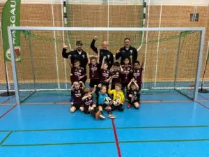 Eine Jugendfußballmannschaft in maroonfarbener Kleidung posiert vor einem Tor in einer Sporthalle.